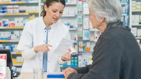 Une pharmacienne explique une notice à une femme âgée, un pilulier bleu posé sur le comptoir